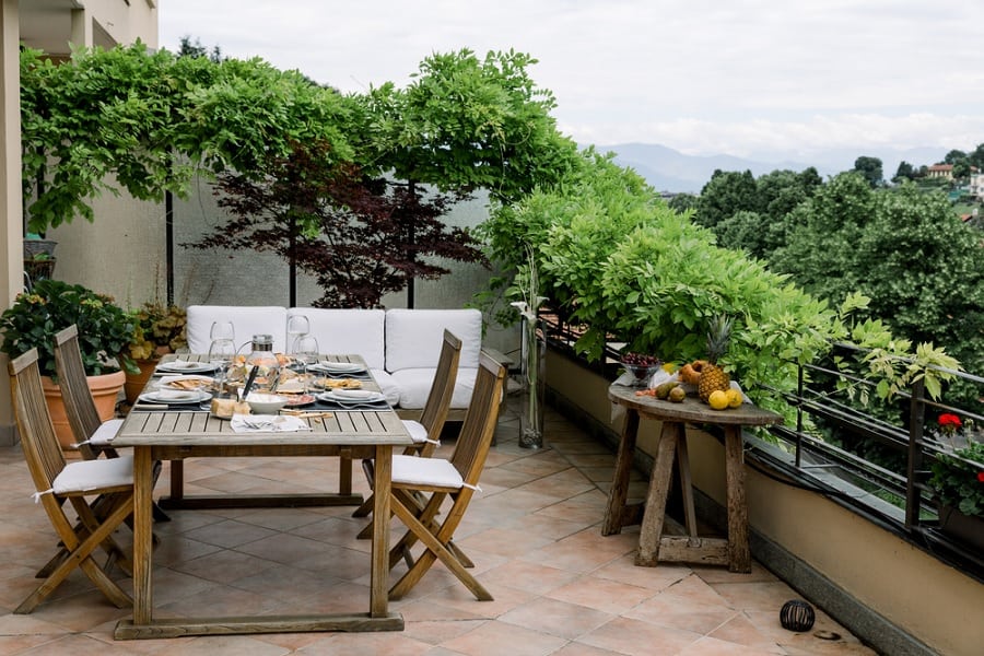 Outdoor patio with dining table, wooden chairs, and lush green plants on a tiled balcony.