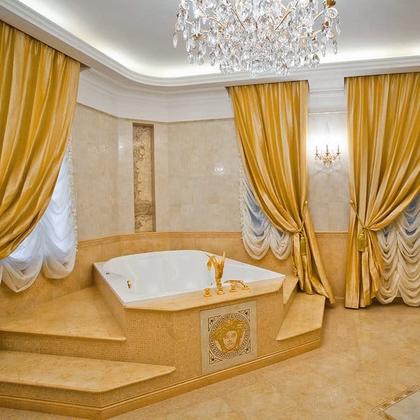 Luxurious bathroom with gold curtains, corner bathtub, and crystal chandelier.