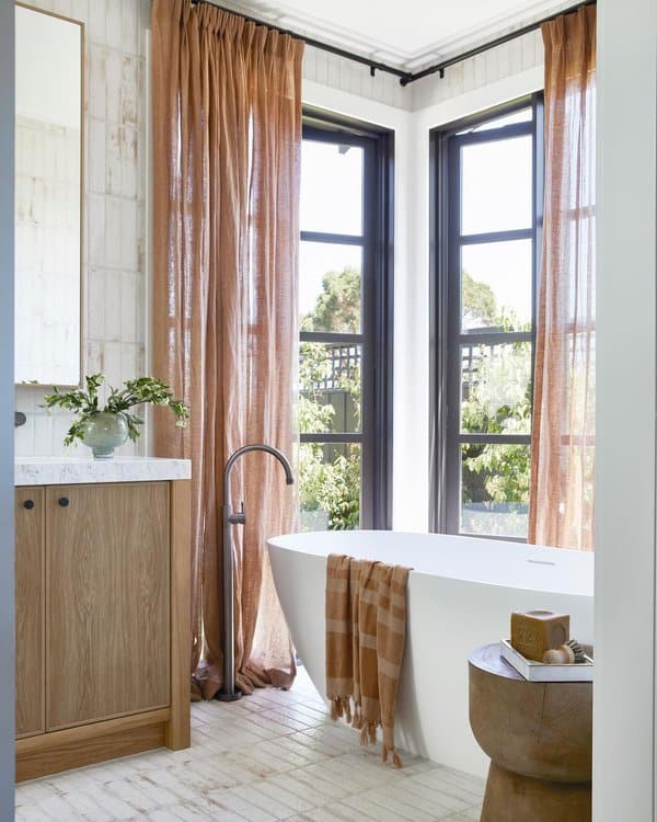 Modern bathroom with sheer brown curtains, freestanding tub, and wood accents.