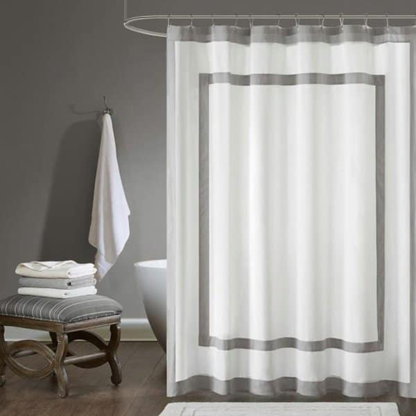 White shower curtain with gray border in a modern bathroom with a stool, towels, and bathtub.