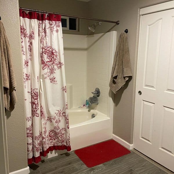 Small bathroom with red and white floral shower curtain, gray walls, and red bath mat.