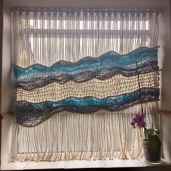 Handwoven macrame curtain with blue and white waves in a small bathroom window.