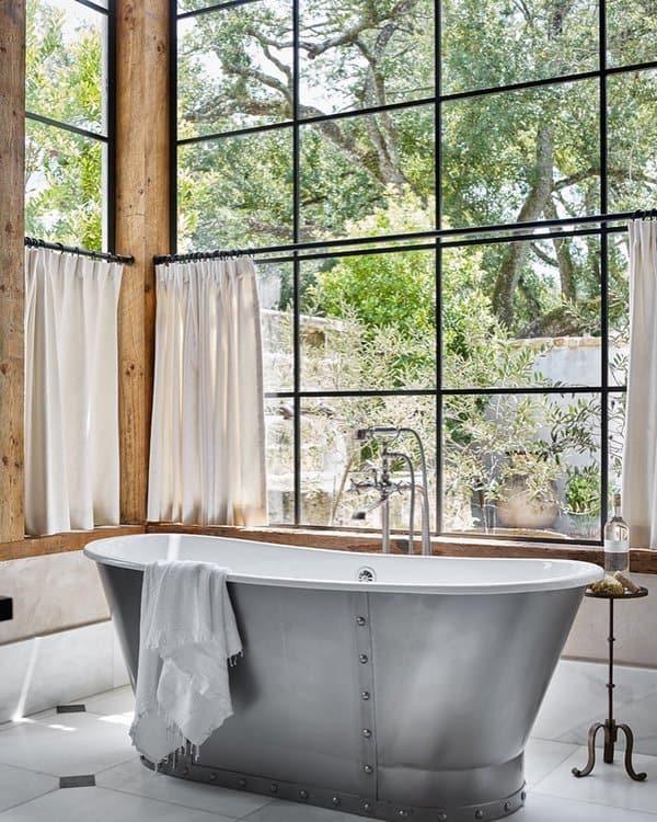 Industrial-style bathroom with large windows, white curtains, and a freestanding metal tub.