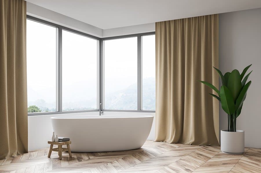 Modern bathroom with freestanding tub, large windows, beige curtains, and wooden floor.