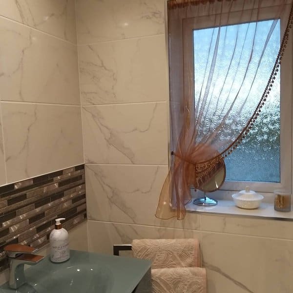 Small bathroom with marble tiles, frosted window, and sheer brown curtain tied to the side.