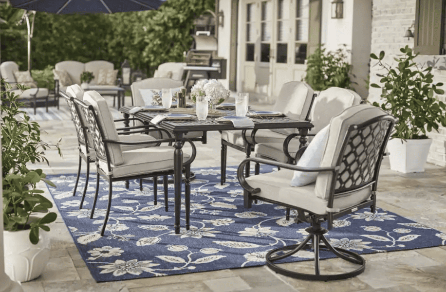 Outdoor dining set with cushioned chairs, a black metal table, and a floral blue rug on a patio.