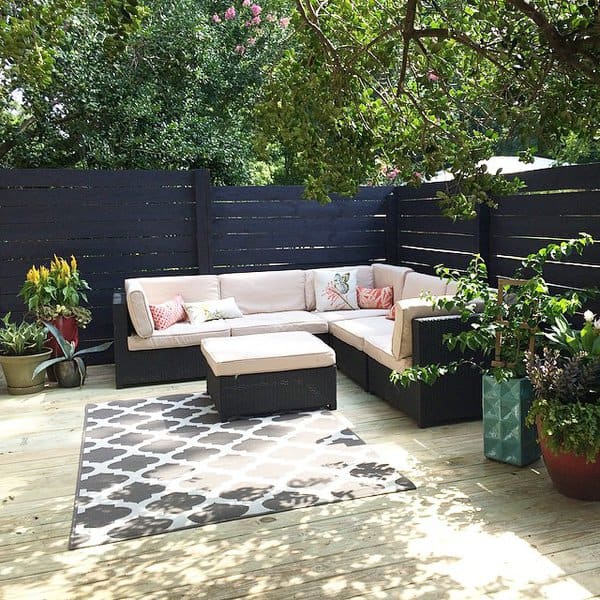 Cozy outdoor deck featuring a black sectional with beige cushions, patterned throw pillows, potted plants, and a stylish area rug against a modern dark wood privacy fence