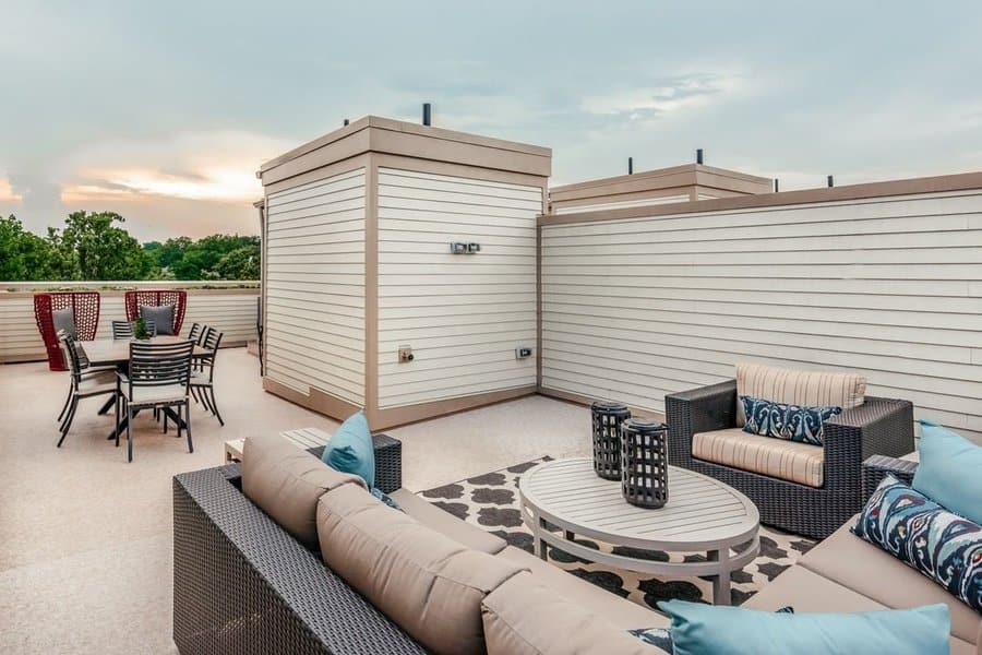 Modern rooftop patio featuring a wicker outdoor seating set with striped and patterned cushions, a stylish round coffee table, decorative lanterns, and a separate dining area with contemporary chairs, all enclosed by light-toned privacy walls