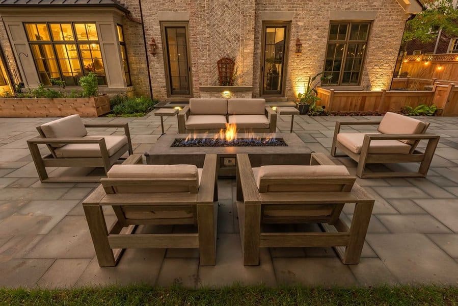 Charming outdoor patio with wooden armchairs and sofas surrounding a modern rectangular fire pit, illuminated by soft ambient lighting against a rustic brick backdrop