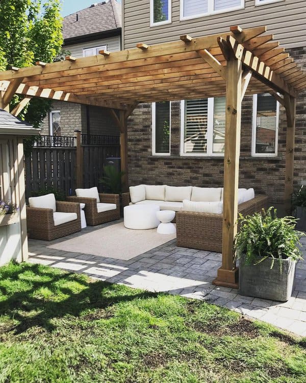 Charming pergola-covered outdoor lounge with wicker seating, plush white cushions, a neutral area rug, and pavers, surrounded by greenery for a cozy backyard retreat