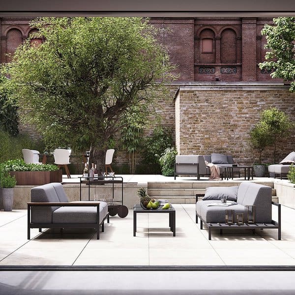 Modern patio with sleek gray seating, black-framed tables, and a lush tree, framed by a rustic brick wall and layered seating areas for a sophisticated outdoor ambiance