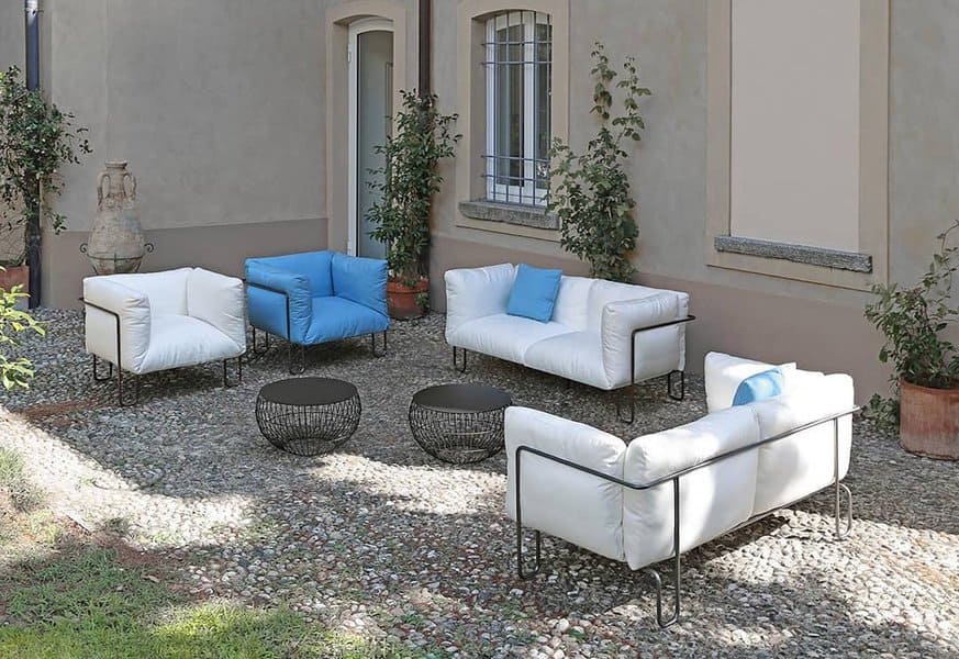 Modern outdoor lounge area featuring sleek white sofas with black metal frames, accented by blue cushions and surrounded by greenery on a pebblestone patio