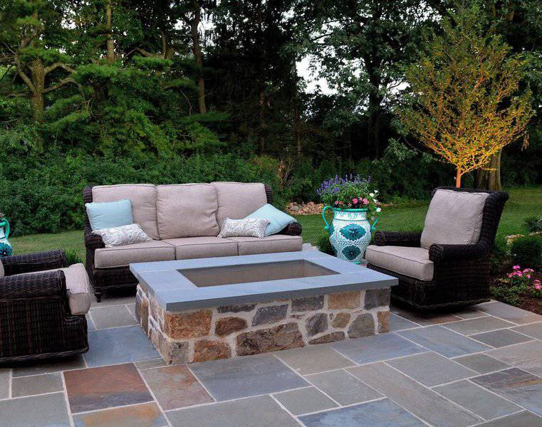 Elegant outdoor seating area with a stone firepit, cozy wicker furniture, and a lush garden backdrop for a tranquil retreat