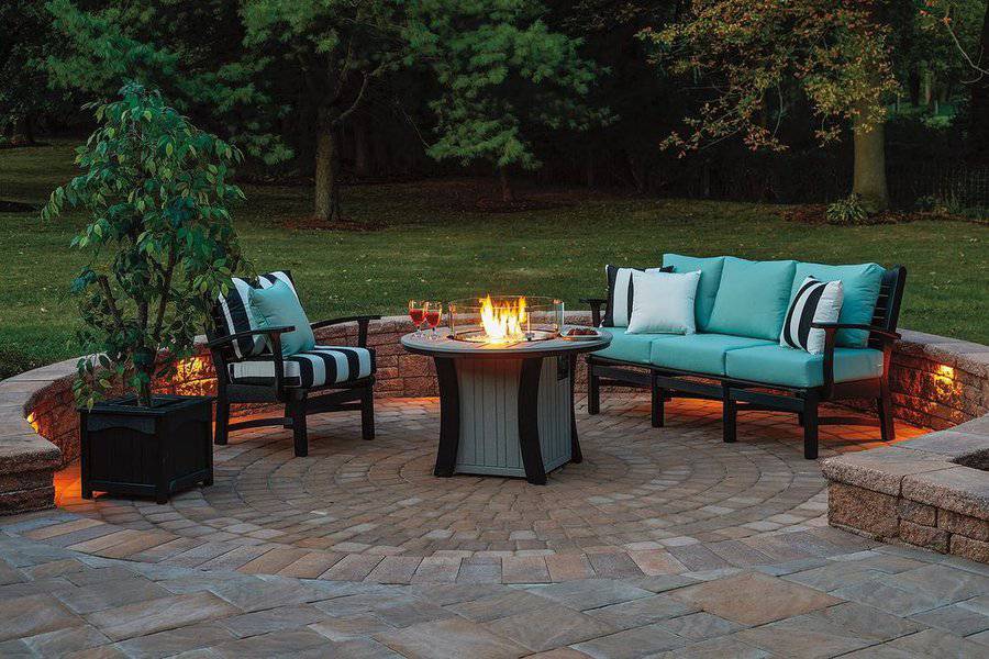 Cozy outdoor seating area with turquoise cushions and striped pillows, centered around a round firepit on a circular paver patio, accented by warm lighting and surrounded by lush greenery.