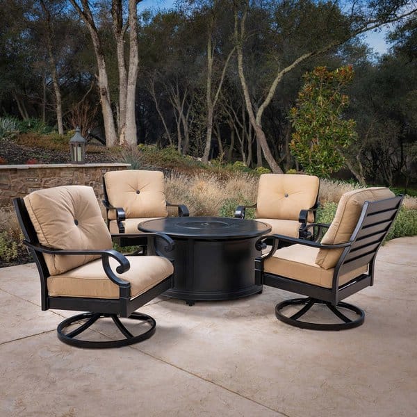 Outdoor seating area featuring four cushioned swivel chairs with beige upholstery, surrounding a sleek black round firepit, set on a stone patio with natural greenery and trees in the background