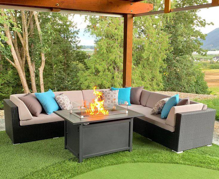 Cozy outdoor seating arrangement featuring an L-shaped wicker sectional sofa with beige cushions and vibrant blue and patterned accent pillows, centered around a modern black firepit table