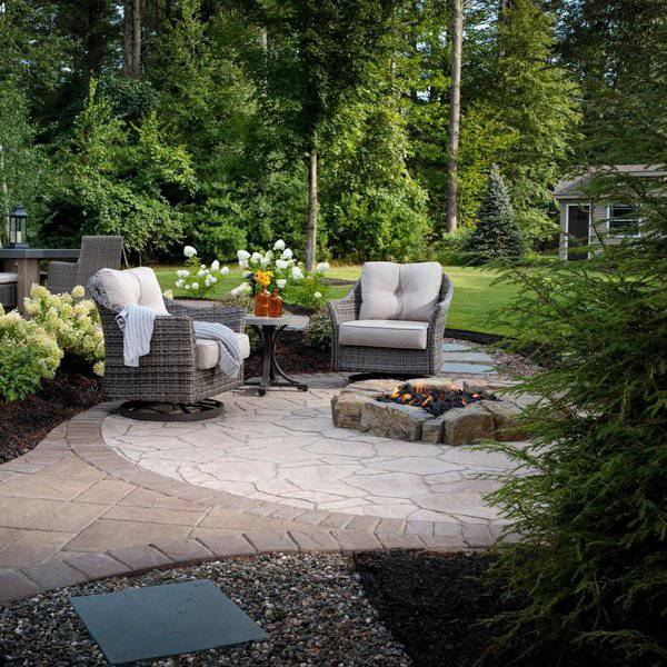 Cozy patio with wicker chairs, stone firepit, lush greenery, and flowers, perfect for relaxing in a serene outdoor setting