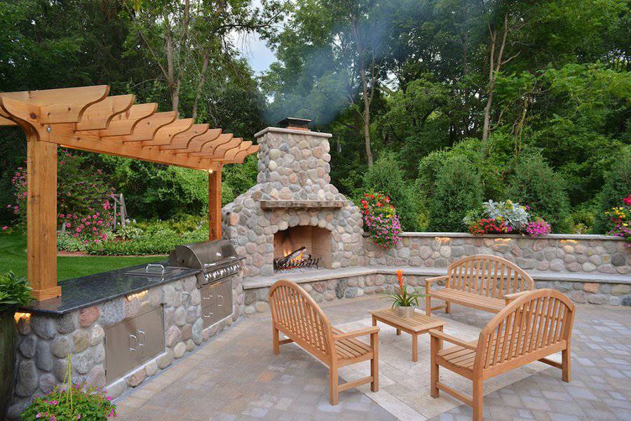 Rustic outdoor kitchen with a stone fireplace, pergola, and wooden seating, surrounded by lush greenery and vibrant flowers