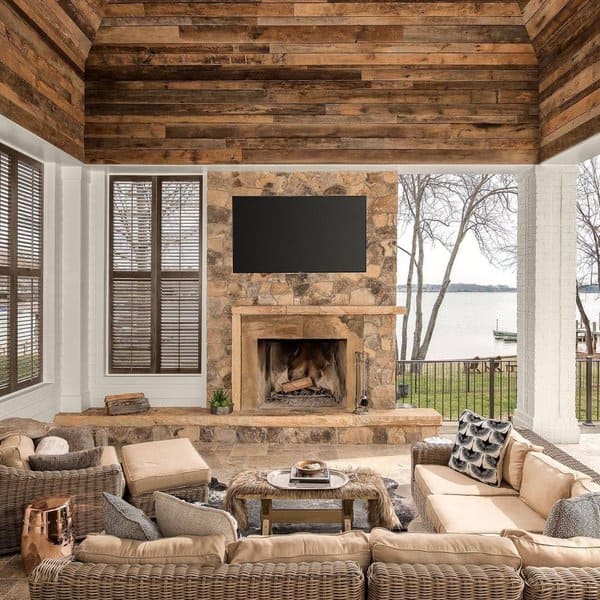 Rustic patio with a stone fireplace, warm wooden ceiling, plush wicker seating, and serene lake views for a tranquil outdoor retreat