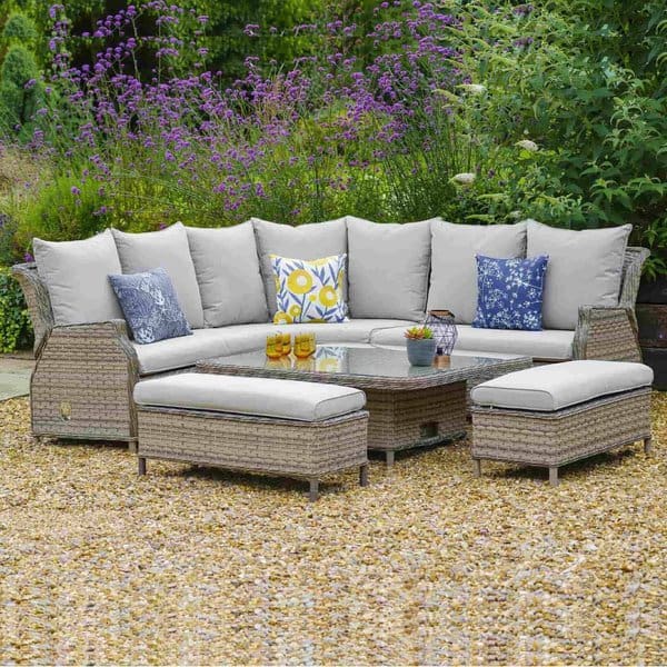 Charming outdoor seating area featuring a cozy wicker sectional sofa with soft beige cushions and colorful floral throw pillows, set against a backdrop of lush greenery and purple flowers