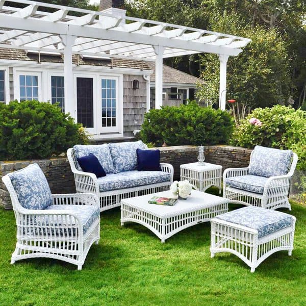 Charming outdoor seating arrangement featuring white wicker furniture with blue floral cushions, set on lush green grass under a white pergola, complementing a cozy cottage backdrop