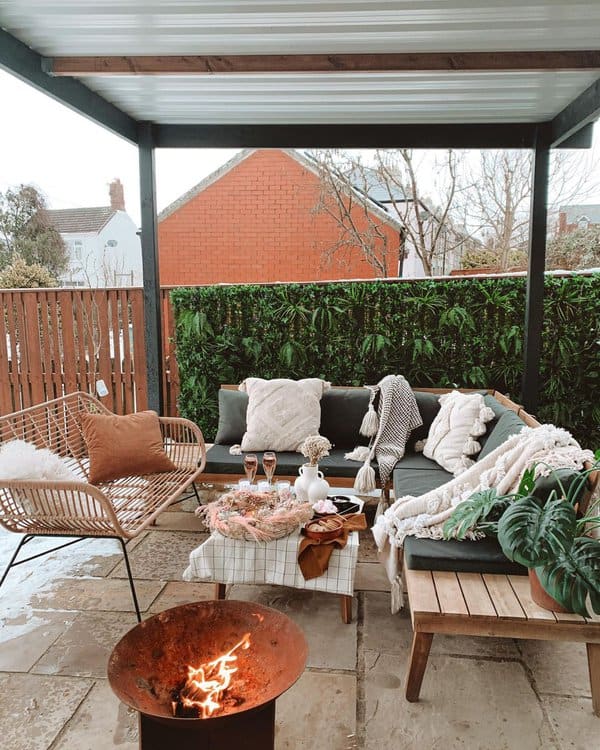 Cozy outdoor space with layered blankets, plush pillows, a wooden coffee table set for a gathering, and a firepit centerpiece, creating a warm and inviting atmosphere