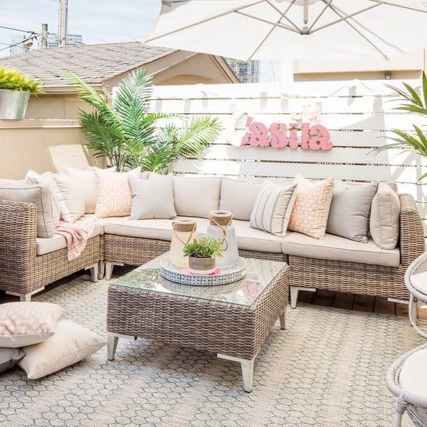 Bright and cheerful outdoor seating area featuring a wicker sectional sofa with pastel cushions, a glass-top coffee table, and lush green plants, complemented by a white privacy fence and umbrella for shade