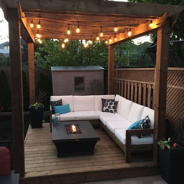 Cozy outdoor deck with a wooden pergola adorned with string lights, featuring a white sectional sofa, decorative cushions, and a firepit table for warmth and ambiance