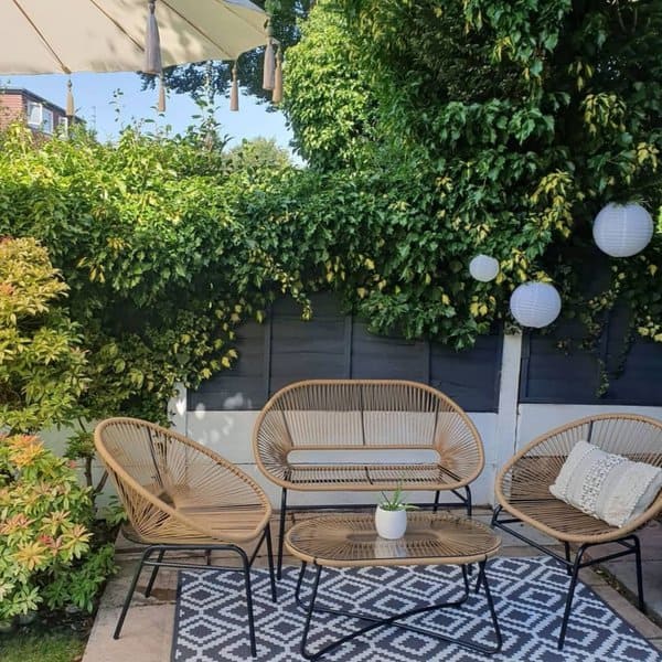 Charming outdoor nook featuring wicker seating with a loveseat and matching chairs, a woven coffee table, a patterned outdoor rug, and lush green foliage as a backdrop