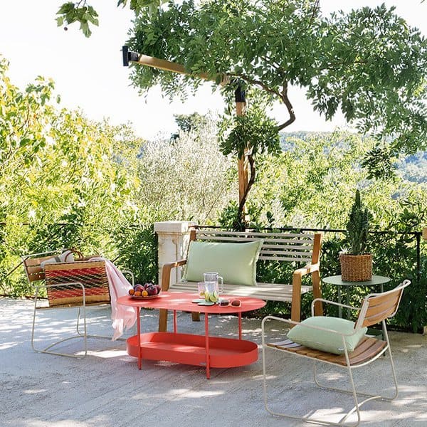 Vibrant outdoor seating area with a red table, light wooden furniture, pastel cushions, and lush greenery creating a picturesque garden view