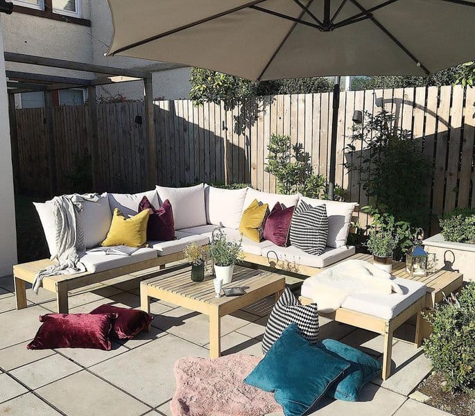 Cozy patio setup with a wooden sectional sofa adorned with colorful cushions, a central coffee table, and soft throw blankets under a large patio umbrella, surrounded by lush greenery and a wooden fence