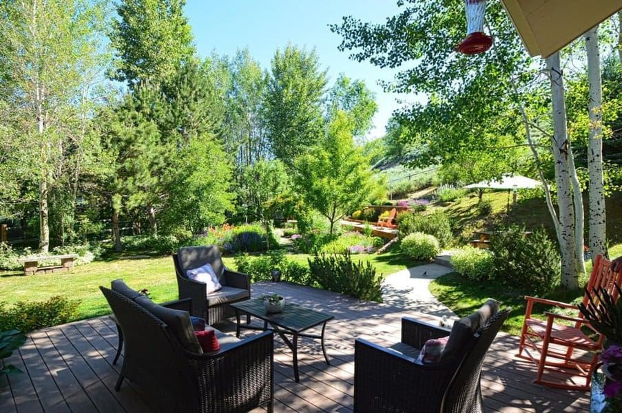 Expansive backyard patio featuring wicker furniture with cozy cushions, overlooking a lush garden filled with trees, flowers, and a serene pathway winding through the vibrant landscape