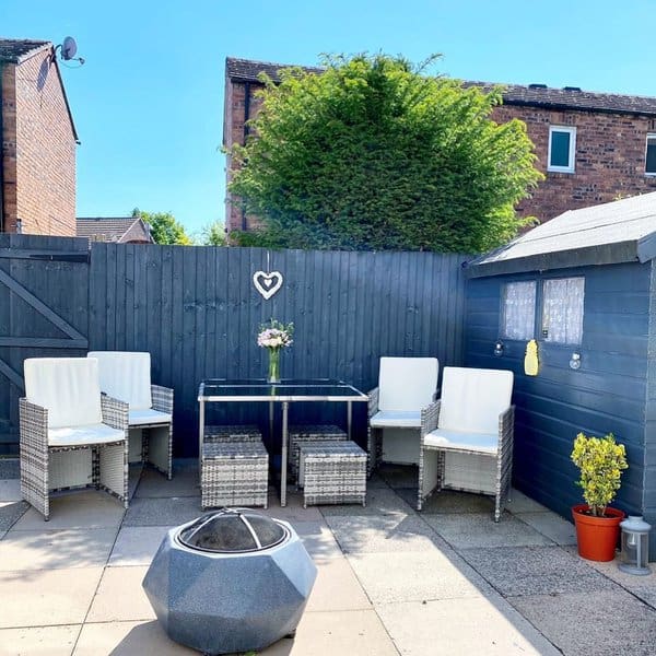 Cozy and minimalist patio setup with wicker furniture and a glass-top table, accented by a stylish firepit and a heart-shaped wall decor, perfect for intimate outdoor gatherings