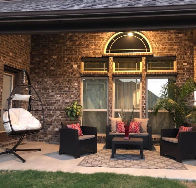 Cozy covered patio with wicker furniture, red accent pillows, a hanging egg chair, and warm lighting against a brick wall backdrop