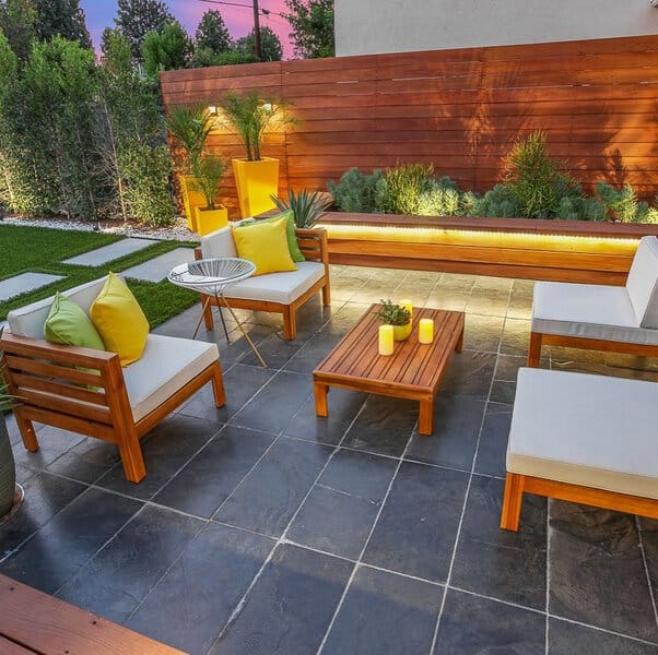 Modern outdoor patio with wooden furniture, vibrant yellow and green cushions, slate flooring, and a sleek wooden privacy fence