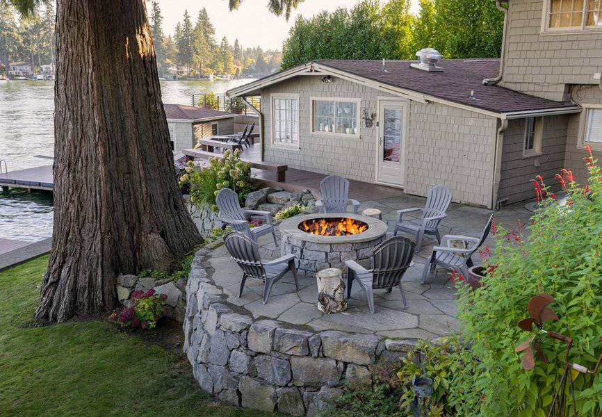 Charming lakeside patio with a stone firepit surrounded by Adirondack chairs, nestled under a large tree with serene water views