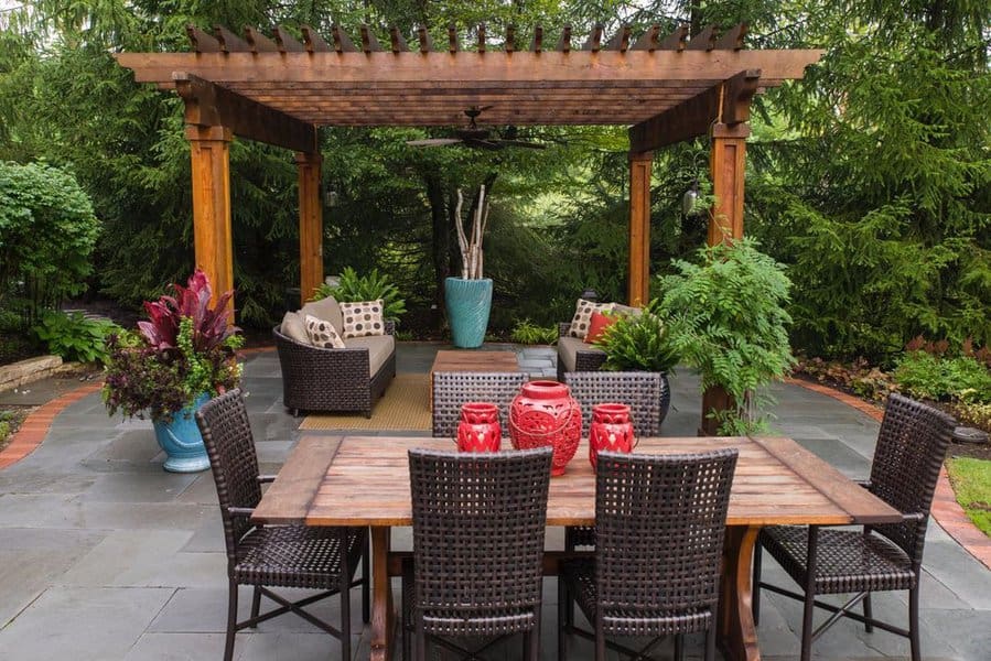 Elegant outdoor space with a wooden pergola, wicker seating, lush greenery, and a dining table adorned with vibrant red accents