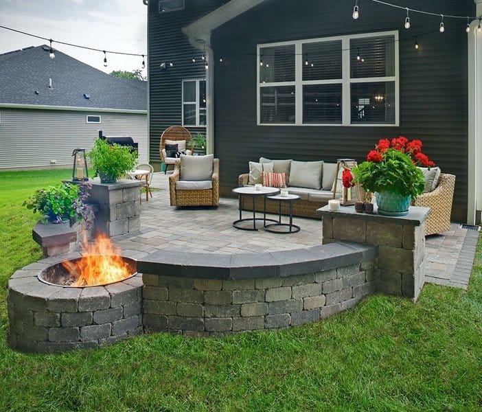 Charming backyard patio with a fire pit, cozy wicker furniture, vibrant red flowers, and string lights creating a warm ambiance