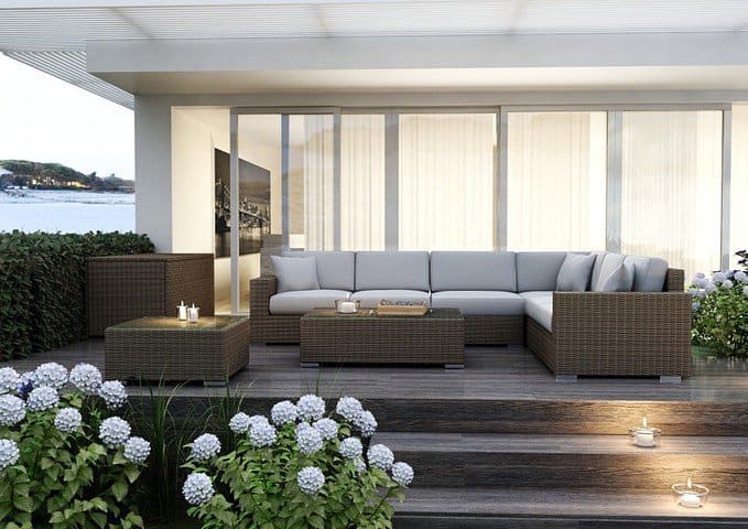 Modern outdoor seating area featuring a sleek wicker sectional sofa with gray cushions, surrounded by blooming white hydrangeas and soft candlelight, overlooking a serene waterfront view