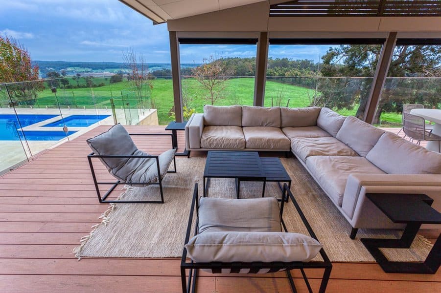 Luxurios outdoor deck with a cozy sectional sofa, sleek black chairs, and glass railings overlooking lush greenery and a pool