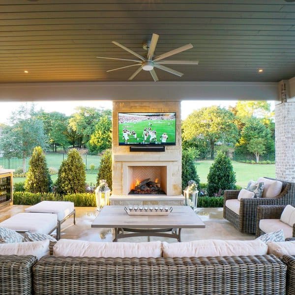 Elegant outdoor patio with a stone fireplace, mounted TV, wicker seating, and lush greenery in a cozy entertainment space