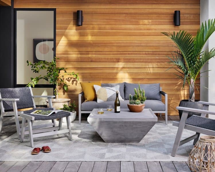 Modern outdoor lounge with gray furniture, geometric concrete table, and wooden panel wall, accented by lush greenery and cozy decor